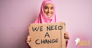 Smiling woman wearing hijab against pink-grey background holding sign saying we need a change
