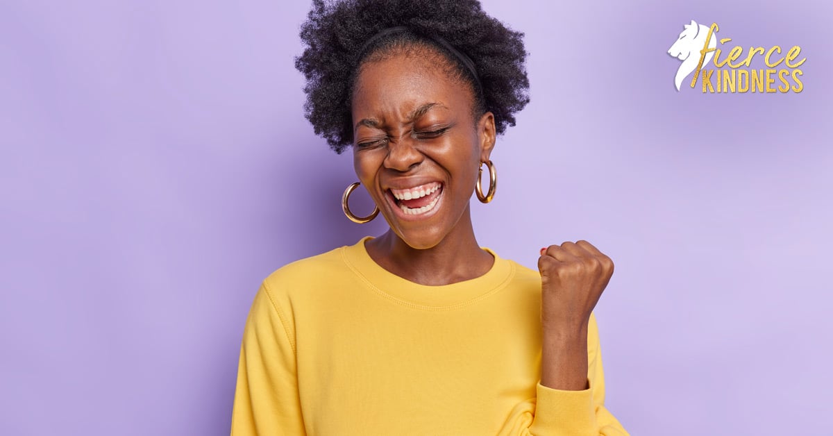Woman with raised fist saying Yes in yellow top against lilac background